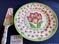 Cake server and matching plate together on dark blue background. Server decorated with pink and red flower and green stem at handle. Plate features large pink tulip in center, surrounded by pink petals, with green leaf and stripe border.
