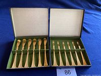 Box with 6 cocktail butter spreaders on left and 6 cocktail lemon forks on right against blue background