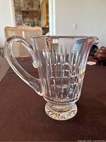 Front and side views of Waterford crystal pitcher on brown table background. Shows clear etched vertical and horizontal lines pattern, solid handle, starburst base, and wide spout.