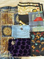 Photo showing six women’s scarves laid flat on a surface. Includes leopard print bordered scarf in gold, black, and brown tones; black, white, and gold chain print scarf; abstract art style scarf with muted greens and reds; butterfly pattern scarf on black background; light blue fringed pashmina; purple floral velvet pattern scarf.