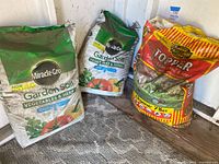 three soil bags leaning against wall: two Miracle-Gro Garden Soil Vegetables & Herbs, one Kellogg Topper Soil for Lawn, Sod & Seed