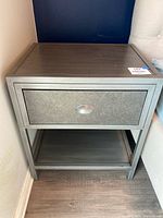 Front view of brushed grey metal nightstand showing single drawer, oval pull, lower shelf