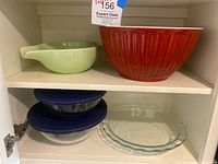 Cabinet shelf with light green serving bowl, red melamine bowl, two Pyrex glass bowls with blue lids stacked, Pyrex quiche dish