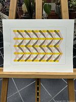 Full view of signed risograph print on paper displayed on an easel, showing colorful geometric chevron pattern in yellow, pink, and brown.