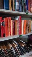 Photo of middle bookshelf with approximately 20+ used paperback fiction books in varied colors, mainly reds and oranges, showing signs of wear.