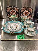 Full set shown with square plates, round plates, cups, saucers, sugar bowl, creamer on stainless steel surface.