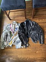 Two vintage leather jackets laid flat on wooden floor. One black classic biker style and one multicolor painted distressed jacket.
