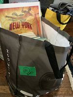 Photo of a grey and white tote bag with handles containing assorted hardcover and paperback books and a New York poster at the back.