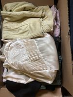Box showing folded assorted clothing including creamy knit sweater and silky embroidered blouse on top