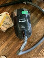 Top angled view of black Eureka The Boss 6830 Series canister vacuum with attached hose and user manual on floor, showing unit label and condition.