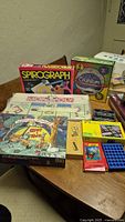 Photo showing a collection of seven board games including Spirograph, Cranium, Monopoly, Oliver & Toes, Domino set, IQ Logic, and Connect Four on a wooden table.