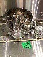 Lot of vintage pewter and silverplate servingware displayed on stainless steel surface showing variety of pitchers, mugs, trays, bowls, and ashtrays
