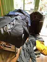 Photo showing various men's jackets including a gray plaid suit jacket, dark blue suit jacket, and a large brown overcoat hanging near a door. Additional clothing items laid on floor include outerwear and sweaters.