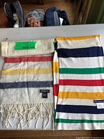 Two Hudson's Bay scarves laid flat on a table, showing classic multicolor stripes in red, yellow, green, and blue on a cream background, one with fringe.