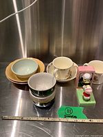 Photo showing grouping of bowls, cups, plates, and a small ceramic figurine on a metal surface with measurement tape in view, also featuring a green label 'Bunnykins'