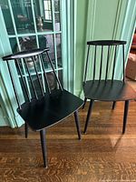 Two black wooden dining chairs shown side by side in front of a green paneled wall, angled view
