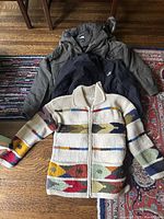 Three men's outerwear items laid on floor: The North Face olive green jacket with hood, black zip-up jacket, and multicolor wool cardigan sweater