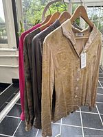 Five silk tops by Windeyer hanging on wooden hangers in natural light, showcasing natural floral hand dyed patterns and colors ranging from cream to pink and grey.