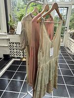 Three silk tops on wooden hangers, hanging on white rack with plants and window in background. Tops include beige/green floral dyed sleeveless with flare hem, dusty rose sleeved top, and another sleeveless pink top.