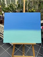 Front view of a rectangular acrylic painting with blue on the upper part fading into green at the bottom, mounted on wooden easel.