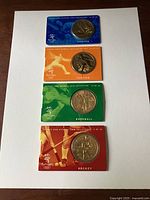 Four coins displayed on white background, each in color-coded card packaging for different sports Sailing, Fencing, Handball, Hockey