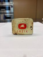 View of front lid of vintage Sheik metal tin with horse and rider logo and 'SHEIK' text, showing wear and rust spots.