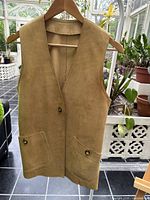 Front view of the tan leather sleeveless vest hanging on a wooden hanger in a bright room with plants.
