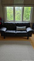 Front view of black leather vintage mid-century modern sofa with one striped throw pillow, situated in front of three-paned window and light hardwood floor.
