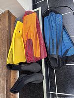 Two pairs of diving fins and two pairs of black water booties shown with yellow and black mesh carrying bags, placed on floor and a wooden bench.