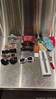 Full assortment of sunglasses, watches, AirPods, Swiss Army multitool, sleep masks, earplugs, and cases arranged on metal surface.