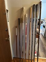 Eight full-sized wooden hockey sticks lined up against a wall, showing varied colors and taped handles/blades. Visible used tape wear and logo branding on sticks.