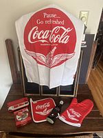 Full lot display showing white Coca-Cola apron with red logo, red oven mitt, pot holder, pen, and 4 coasters on a table.