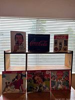 Photo showing three hardcover Coca-Cola books on upper shelf and three Coca-Cola vintage-style calendars on lower shelf in front of window blinds.
