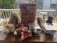 View of all 10 Coca-Cola plush collectibles with the vintage tin container in the background, showing varied sizes and species themed around Coca-Cola.
