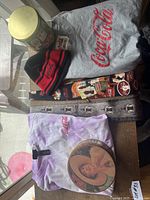 Photo showing the gray T-shirt, vintage Coca-Cola tin can, black/red beanie hat and purple tie-dye T-shirt with vintage Coca-Cola design laid out on a table.