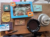Photo of various vintage smoking accessories including tobacco packs, pipe, ashtrays, and Smoking Elephant cigarette dispenser in box.