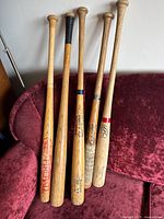 Five vintage wooden baseball bats made by different brands including Louisville Slugger and Rawlings, displayed upright on a red couch.