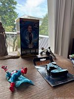 Overview of the boxed Doctor Who Tenth Doctor bobblehead figure, Marvel figure lying on the floor with red gloves and blue outfit, and another figure posing with weapon lying down on the table.