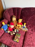 Collection of mixed plush and plastic collectible figures along with a VHS tape laid out on a maroon sofa.