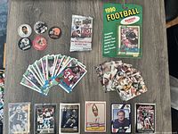 Top-down view showing multiple NFL football trading cards, collegiate cards, football-related pins/buttons, unopened 1990 Topps Raiders football card pack, and loose card stacks.