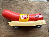 Side view of red hot dog piggy bank with yellow Oscar Mayer sticker on beige bun base with wheels, showing minor wear on surface.