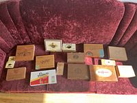 Photo showing 16 assorted empty cigar boxes on a red sofa, wooden and metal boxes with various brand markings including Don Tomas, El Producto, Panter, and others.