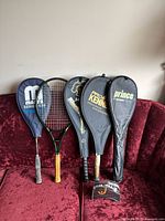 Five rackets standing upright with their respective covers on, showing front views of covers and rackets handles.