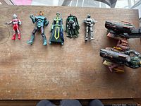 Wide view of five Guyver action figures in a row with two mounted sci-fi guns displayed on a wooden surface.
