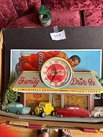 Coca-Cola Family Drive-In clock showing retro diner with vintage cars in front and a large round clock face with Coca-Cola logo at top
