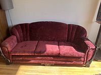 Full front view of the vintage maroon velvet sofa showing the curved and tufted backrest, seat cushions, and wooden carved base and arm on the right side