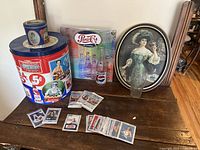 Photo showing large and small vintage Pepsi tins, framed art with a woman holding a glass, Pepsi bottle, glass, and trading cards spread out on a table