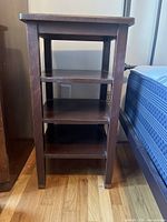 Front view of the nightstand showing three shelves and straight legs on hardwood flooring next to a bed.