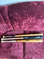Three wooden baseball bats laid on a red velvet couch, showing visible wear, discoloration, and different brand markings.