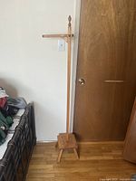 Photo showing full view of wooden vintage coat rack standing next to door and bed frame, displaying its height, natural wood finish, and base with three legs.